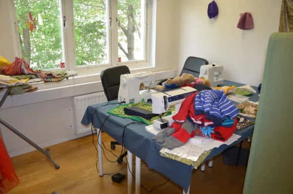 Rörigt syrum med textilier, maskiner och stolar under ett fönster.