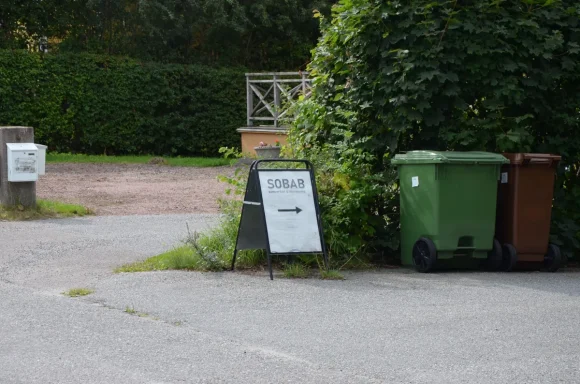 Skylt med text "Återvinning" vid soptunnor i en grönskande omgivning.