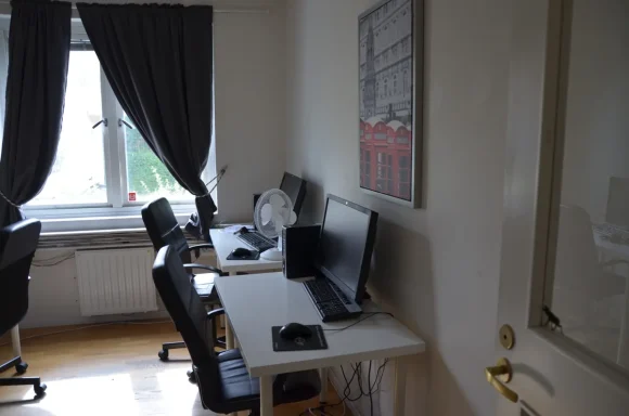 Kontorsutrymme med två skrivbord, datorer och svarta kontorsstolar. Gardiner vid fönstret.